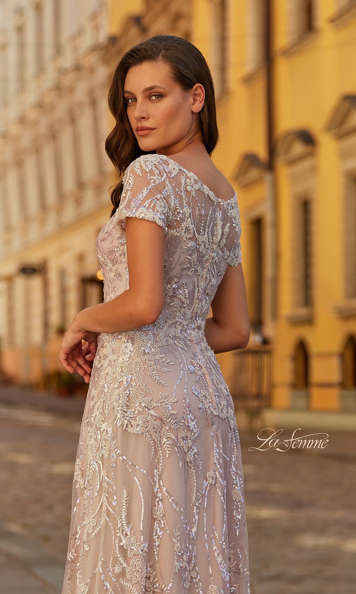 A woman in the La Femme 31838 long formal dress, a light mauve, short-sleeved, floor-length gown with beaded embroidery, stands on a cobblestone street by yellow buildings.